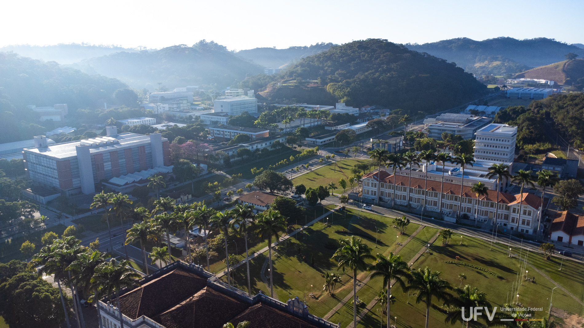 Foto aérea do campus Viçosa da UFV. Vê-se parte da avenida principal, a biblioteca central, palmeiras e um dos alojamentos. 