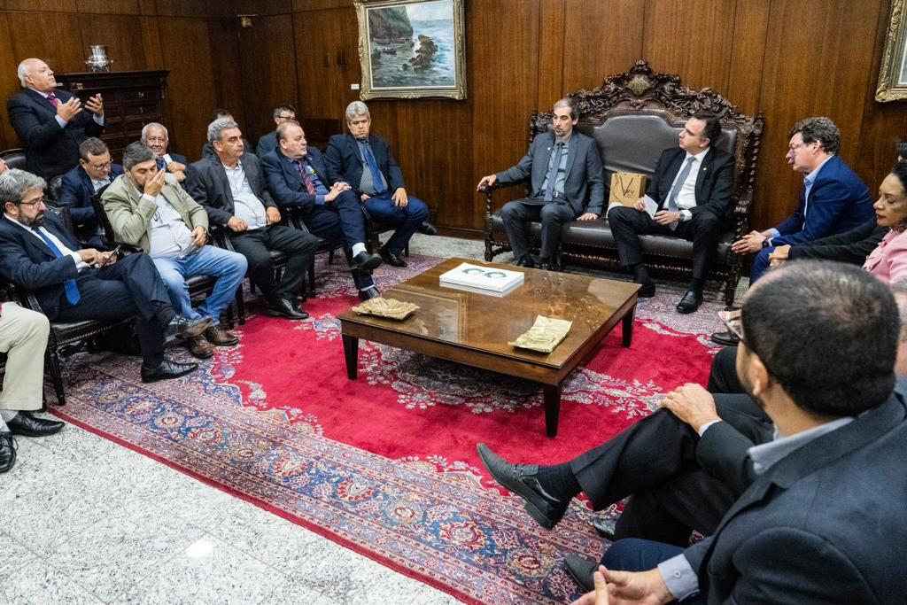 Demetrius, Rodrigo Pacheco e demais participantes sentados em sofás colocados em círculo, numa grande sala. 