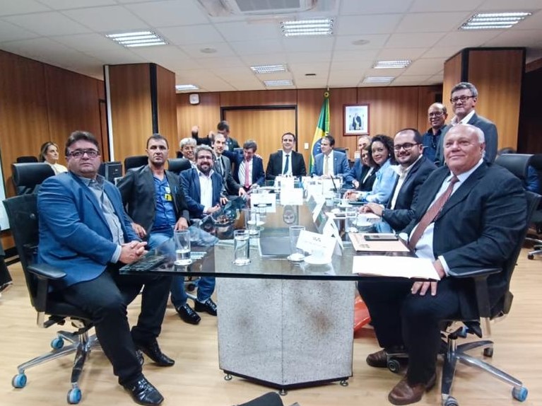 Demetrius, Rodrigo Pacheco e demais participantes ao redor de uma mesa de vidro numa grande sala com paredes de madeira. 