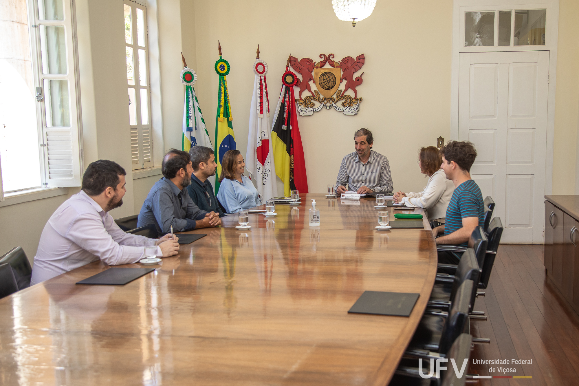 Foto de Demetrius, Rejane e demais participantes de pé, à frente do brasão da U F V. 