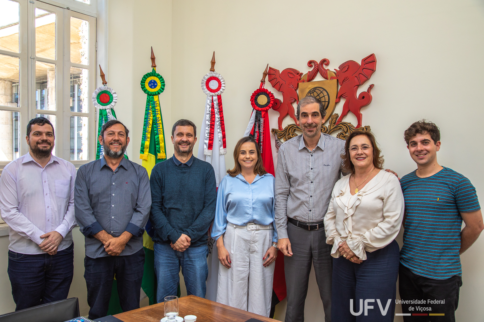 Foto de Demetrius e demais participantes ao redor de uma grande mesa de madeira. Na parede ao fundo, brasão da U F V. 