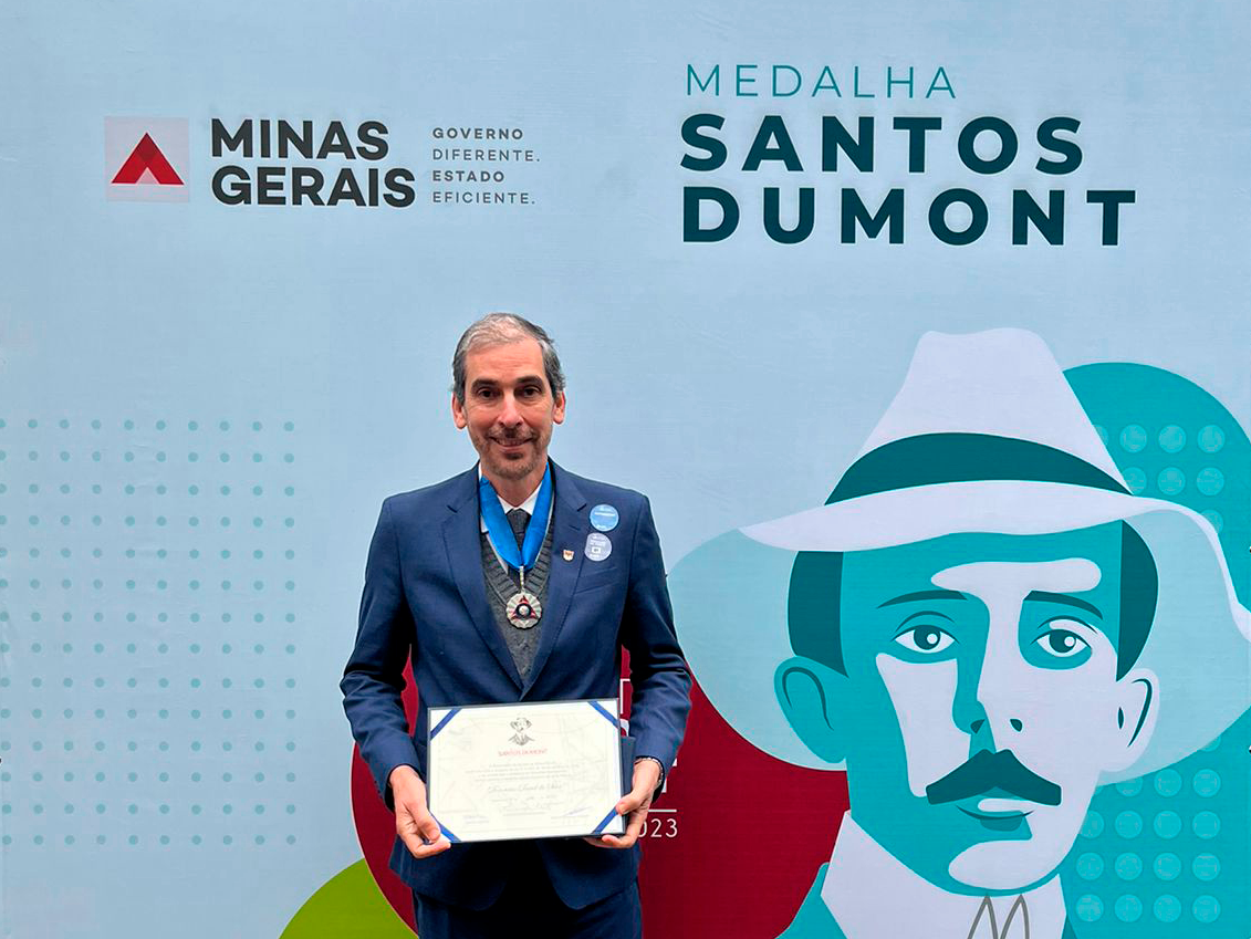 Foto de Demetrius segurando um certificado e usando a Medalha. Atrás dele, painel azul com imagem de Santos Dumont. 