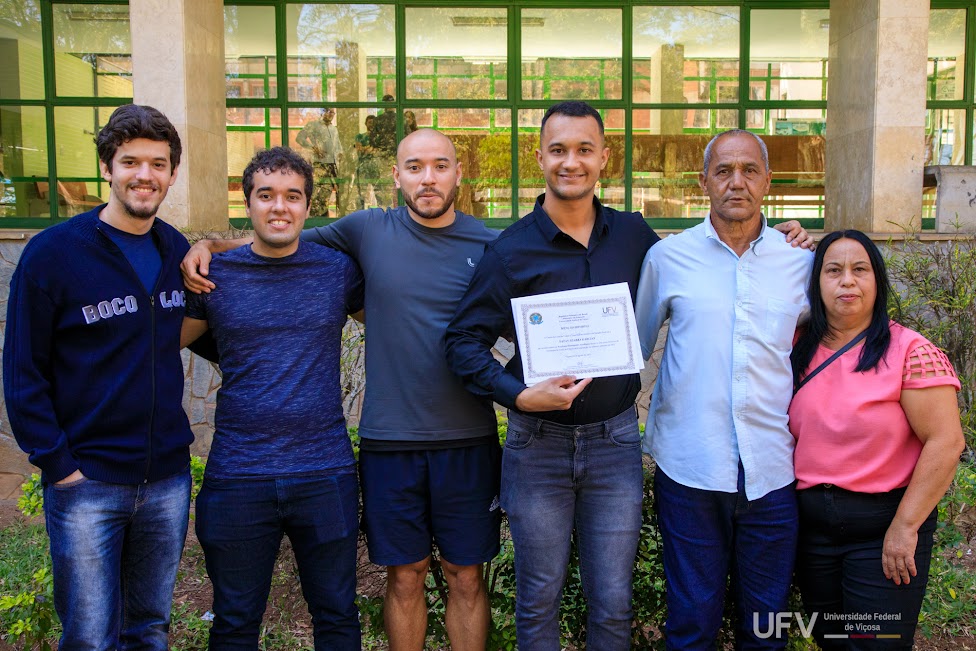 Foto de Nata, seus pais e seus amigos posando para a foto. Natan segura o certificado em frente ao peito. 