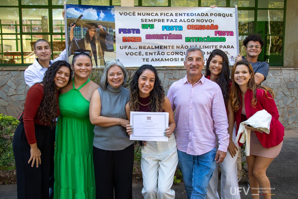 Foto de Luiza, segurando seu certificado junto a seus pais. Atrás deles, amigos seguram uma faixa comemorativa. 