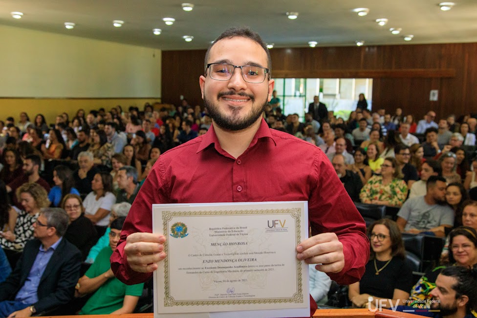 Foto de Enzo logo após receber sua homenagem, em frente a um auditório lotado. Ele segura o certificado em frente ao peito. 