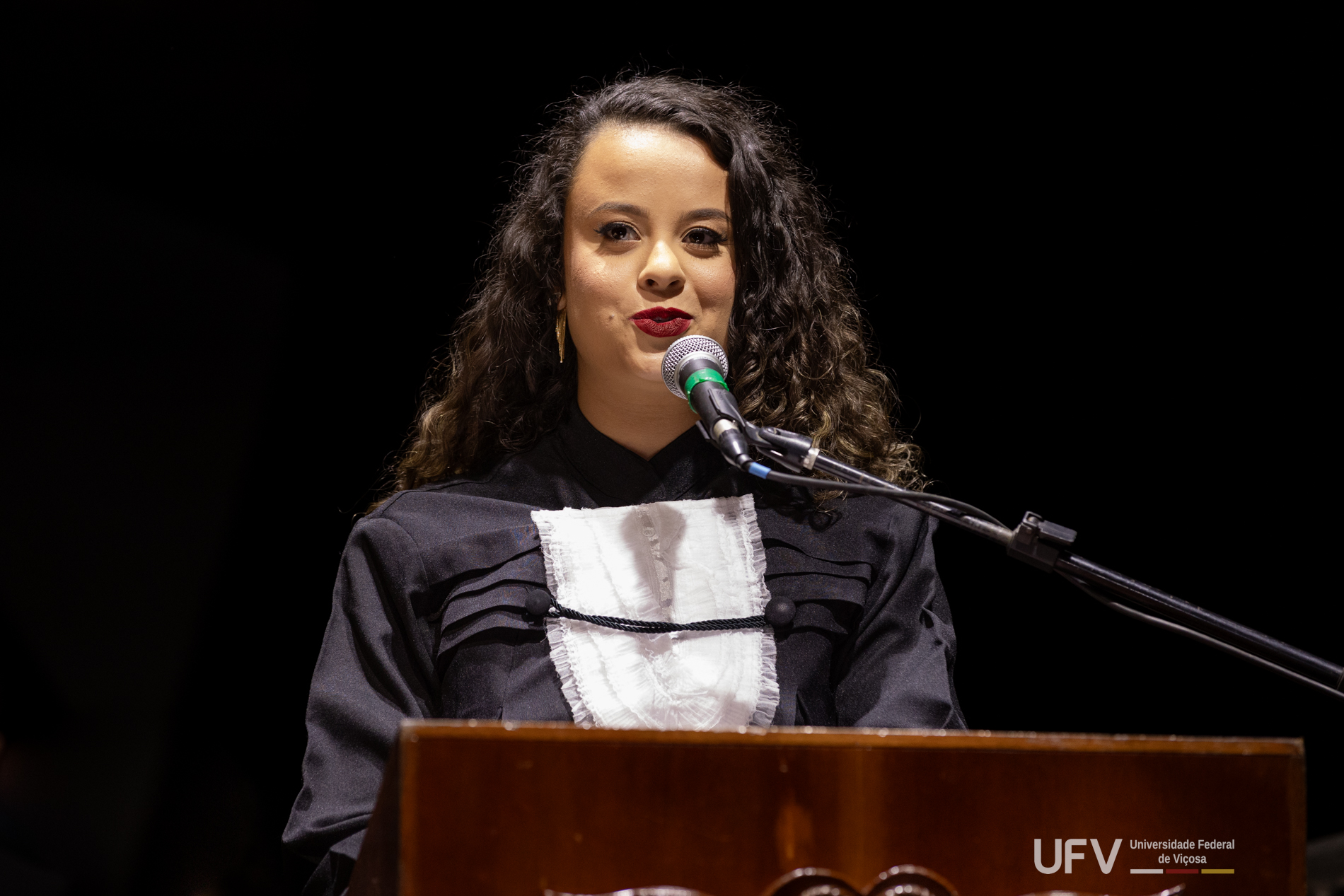 Foto de Maria Luíza, mulher jovem, parda, cabelos pretos longos e ondulados e batom vermelho escuro, durante sua fala. 