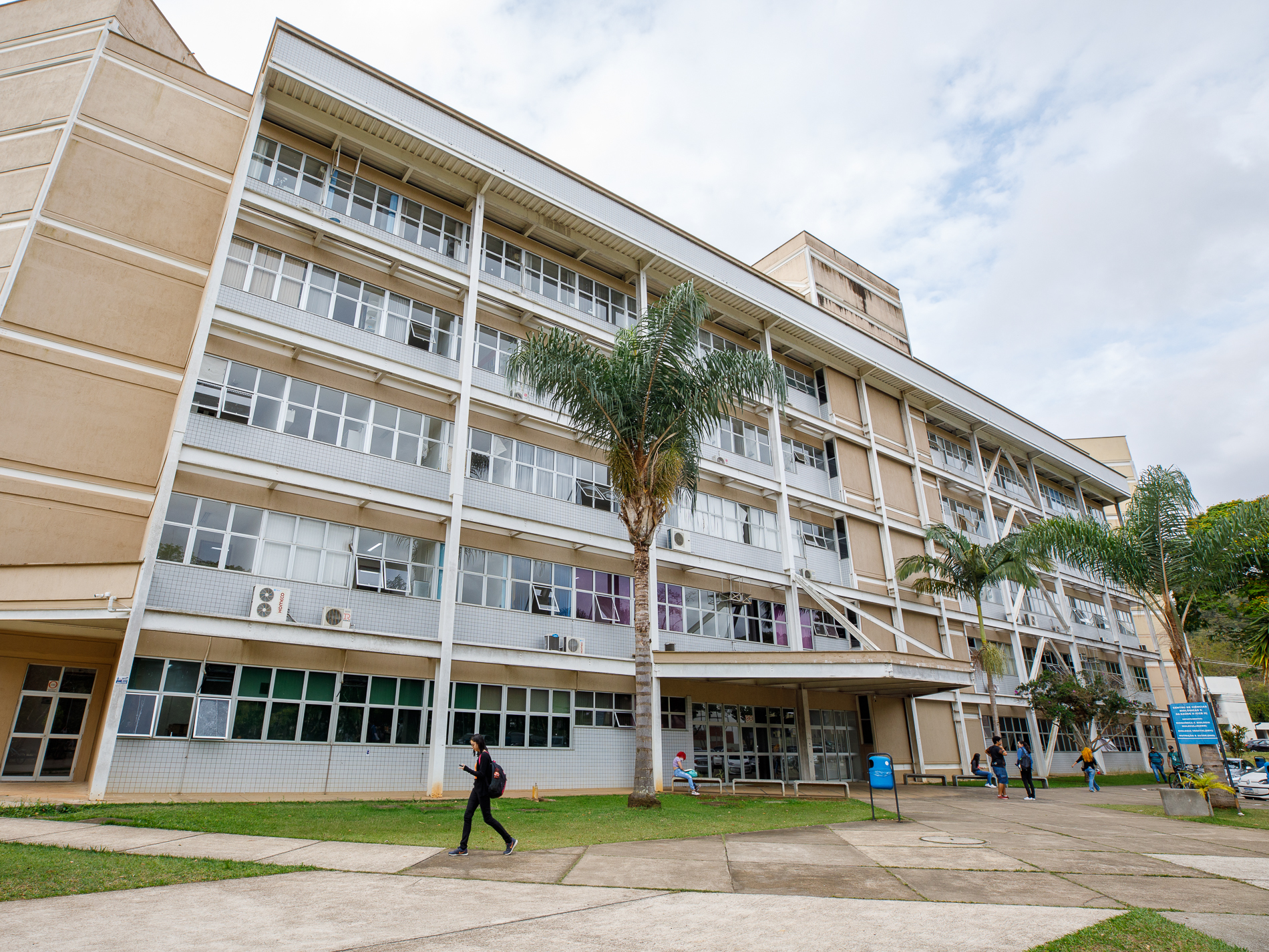 Foto da fachada de um prédio de 4 andares, em cinza e bege, com grandes janelas em vidro e alumínio. 