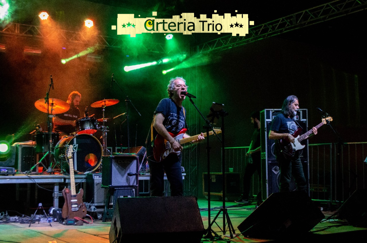 Foto do trio num palco iluminado em verde e vermelho. São: um baterista, um guitarrista/vocal e um baixista/vocal. 