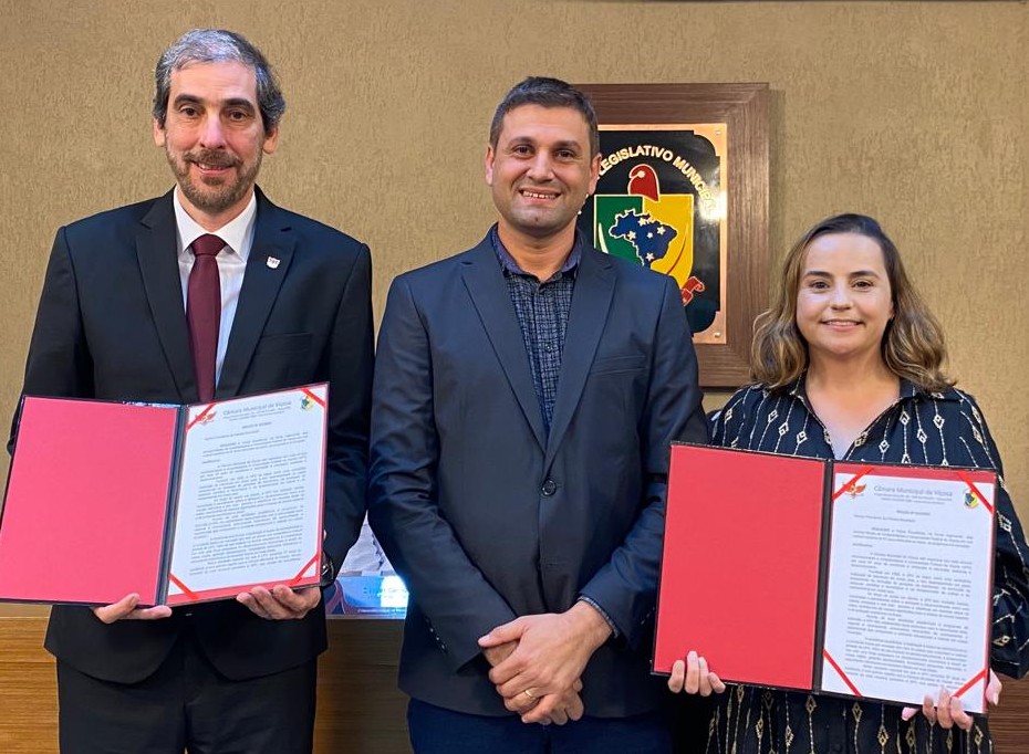 Demetrius e Rejane posam para foto, segurando os certificados de homenagem em frente ao corpo. Marcos está entre os dois. 