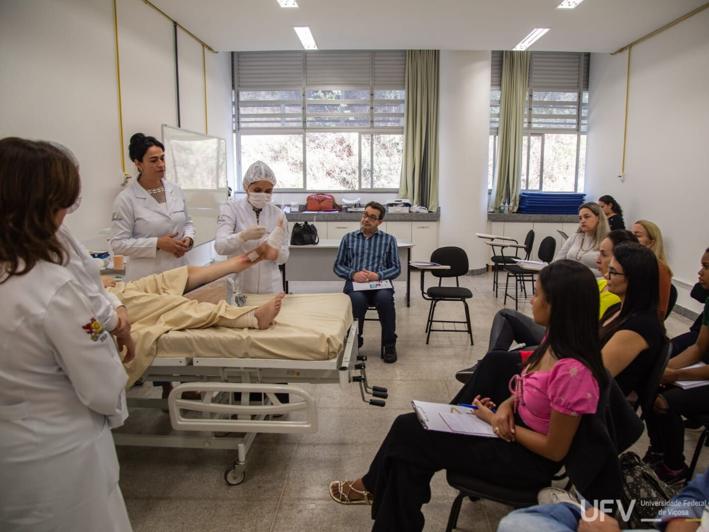 Num ambulatório, enfermeira realiza tratamento de ferimentos em um paciente real, sendo observada por um grupo de pessoas. 