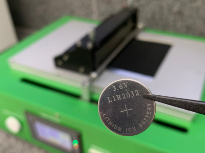 Foto de bateria em formato de moeda sendo segurada no ar por uma pinça. Ao fundo, equipamento eletrônico verde. 