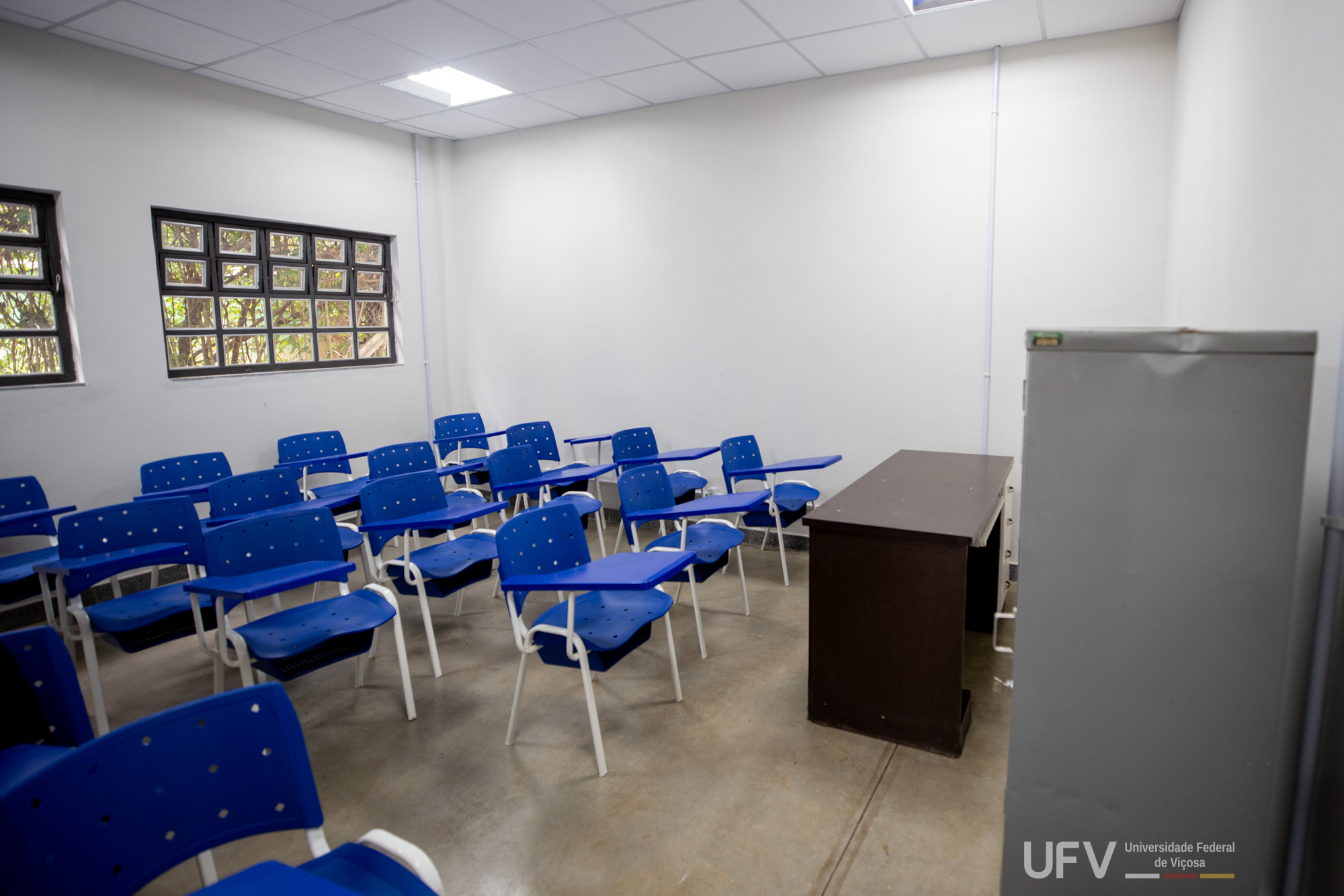 Pequena sala de aula com janelas quadriculadas e cadeiras azuis. No canto direito da foto, uma mesa marrom. 