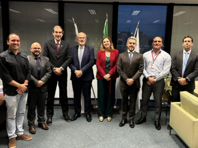 Demetrius e demais autoridades presentes à reunião, de pé lado a lado em frente a uma grande parede de vidro. 