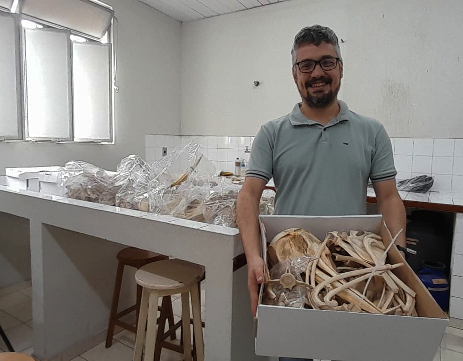 Foto de Guilherme em uma sala com ladrilhos brancos. Ele segura, sorridente, uma grande caixa com ossos variados. 