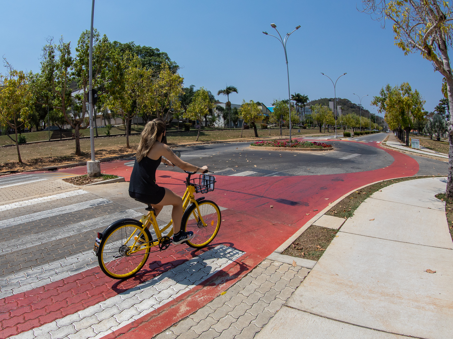 Mulher jovem, branca e loira passeia de bicicleta por uma ciclovia pintada de vermelho na pista, numa grande avenida reta. 