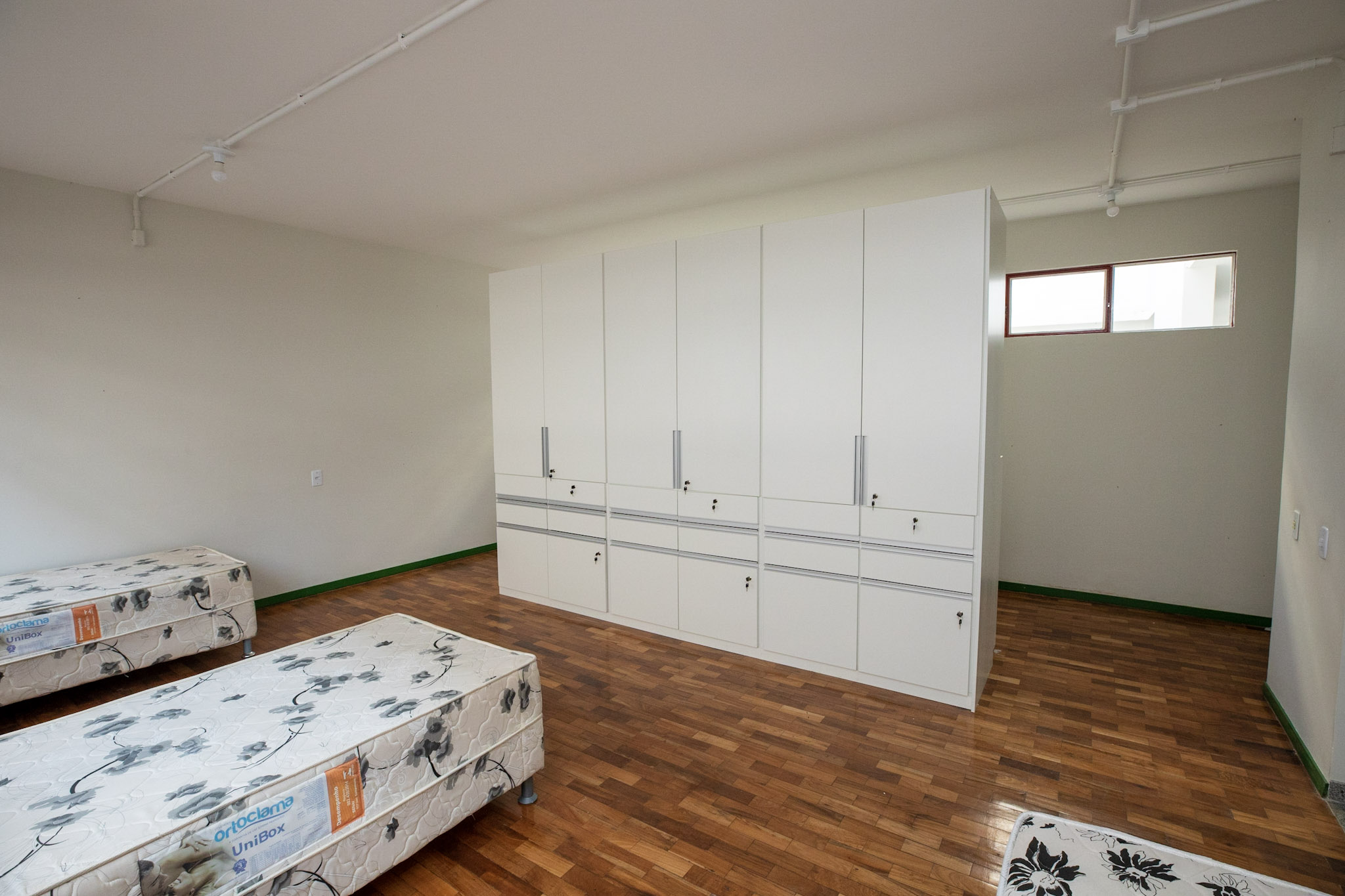 Interior de um quarto de alojamento, com piso de madeira, 3 camas box e um armário branco com portas e gavetas. 