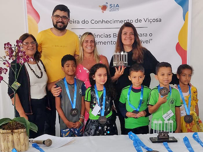 4 adultos e 5 crianças, cada uma com uma medalha, posam para a foto. Uma das mulheres segura um troféu na forma das 4 Pilastras da UFV. 