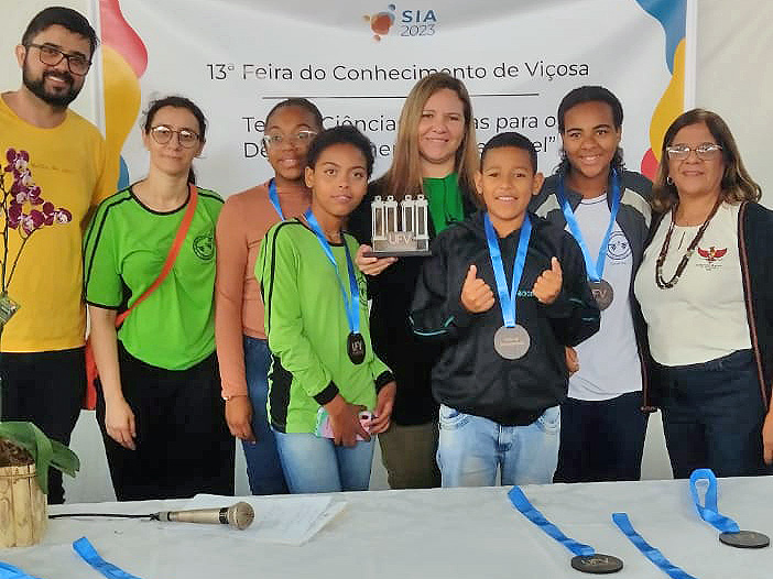 4 adultos e 4 adolescentes, cada um com uma medalha, posam para a foto. Uma das mulheres segura um trofeu na forma das 4 Pilastras da UFV. 