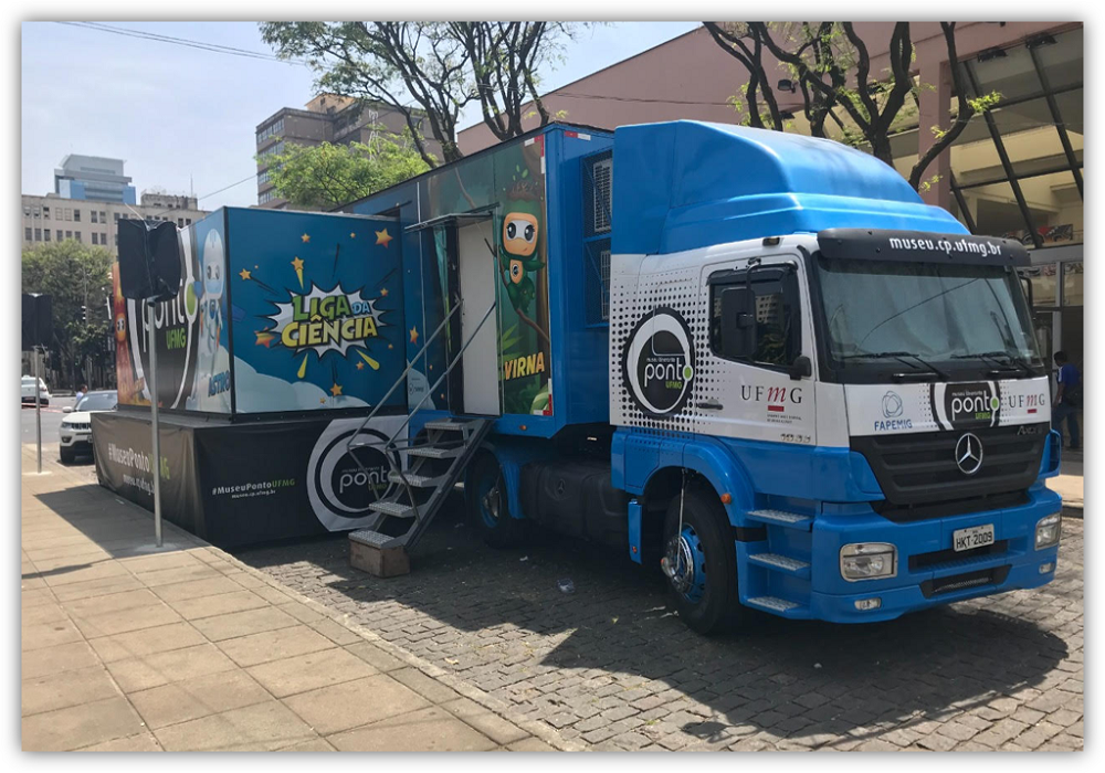 Caminhão azul e branco. Uma escada com corrimão dá acesso ao interior do baú, que tem um baú menor acoplado na lateral. 