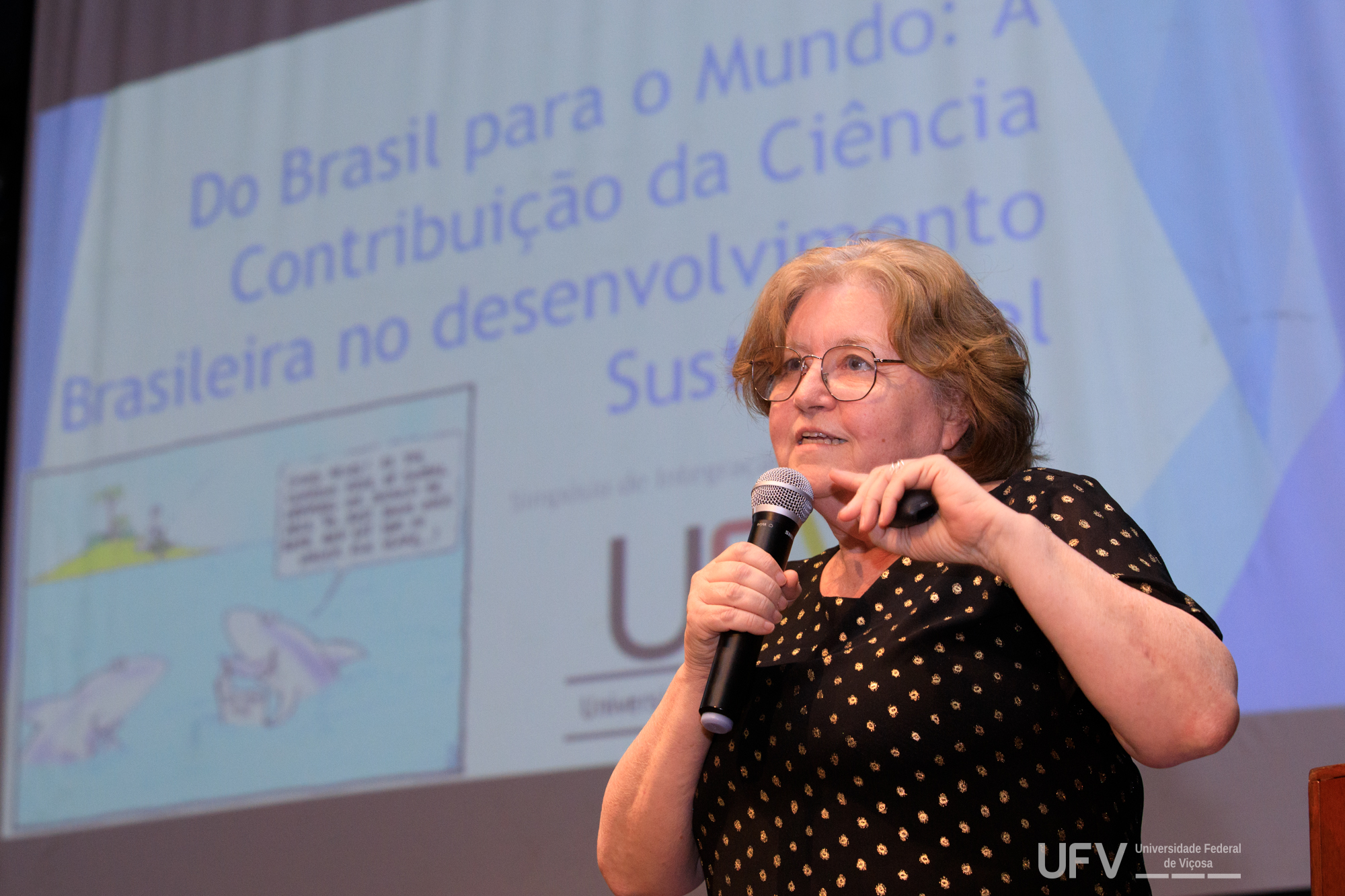 Connie fala ao microfone. Mulher branca, meia-idade, cabelo loiro ondulado e curto, óculos e camisa preta e dourada. 