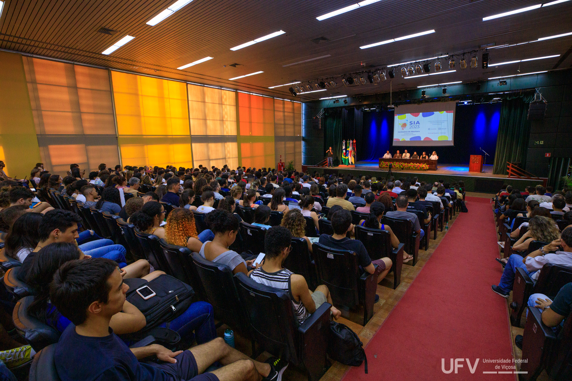 Auditório com jovens em todas as cadeiras. Ao fundo, palco iluminado de azul com uma grande mesa e pessoas sentadas. 