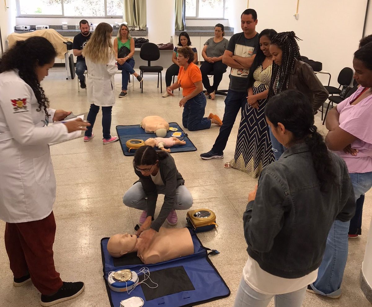Duas mulheres de jaleco estão de pé. No chão, outras duas estão ajoelhadas junto a bonecos de simulação médica. 
