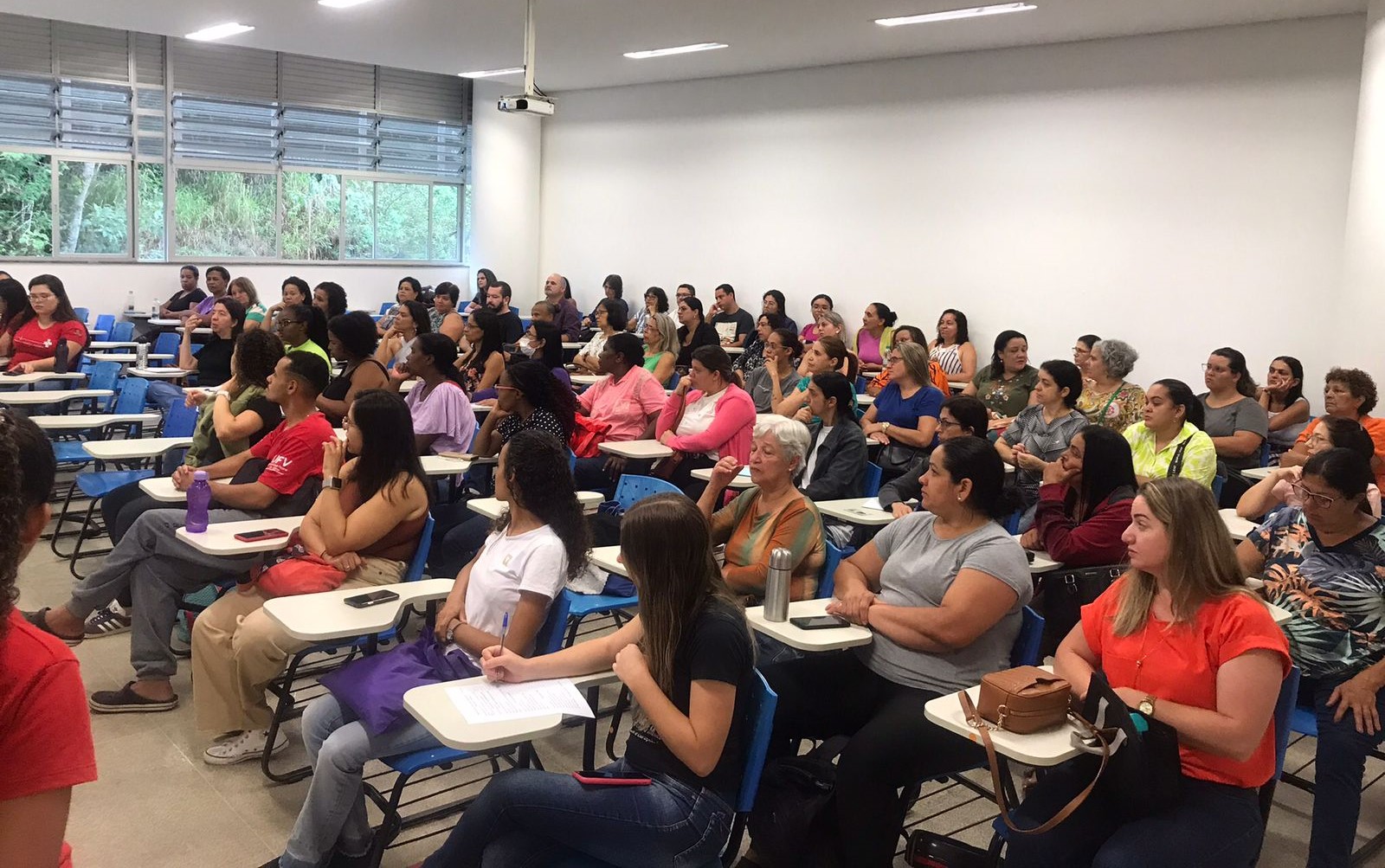 Grande sala de aula com muitas carteiras, todas ocupadas principalmente por mulheres de várias idades. 