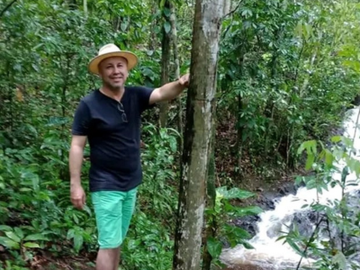 Joenilton está de pé numa área arborizada, apoiado em um tronco. Ele usa um pequeno chapéu de palha e bermuda azul claro. 