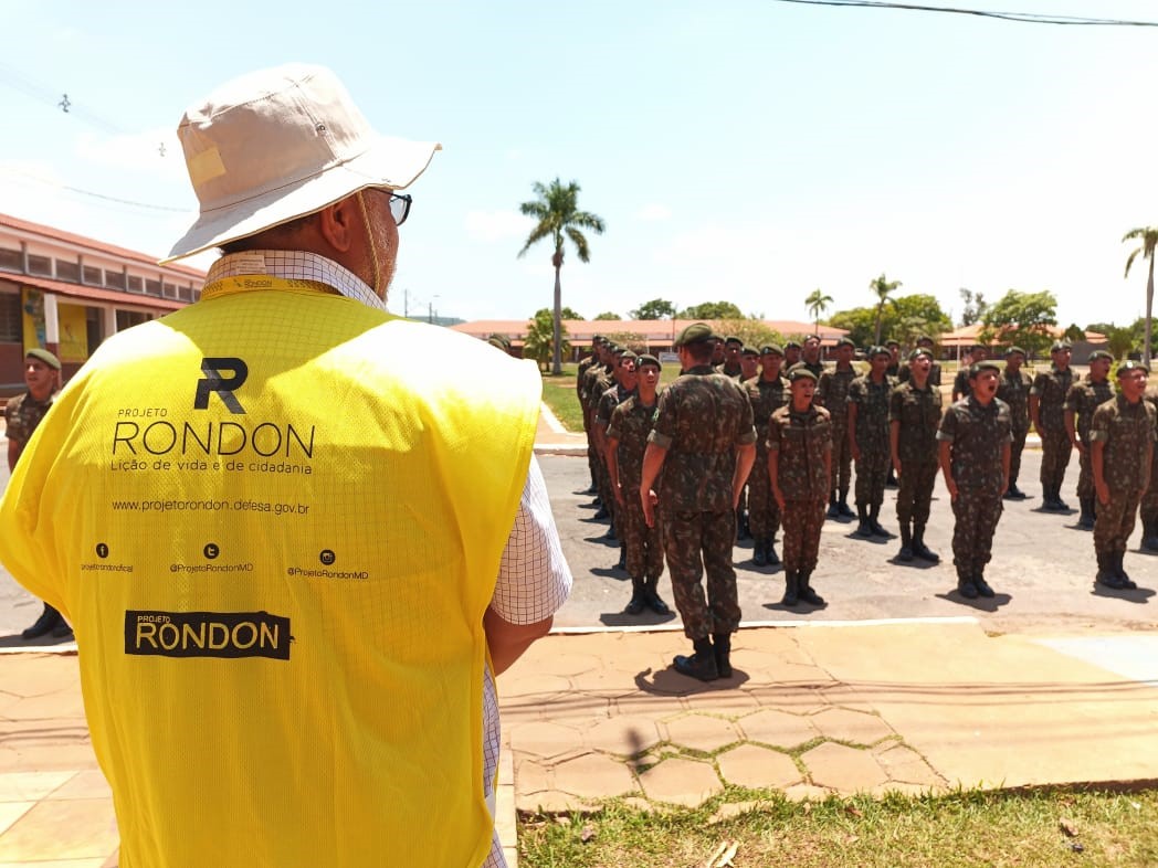 Homem costas, usando colete amarelo e chapéu tipo australiano, observando um grupo de militares em treinamento. 