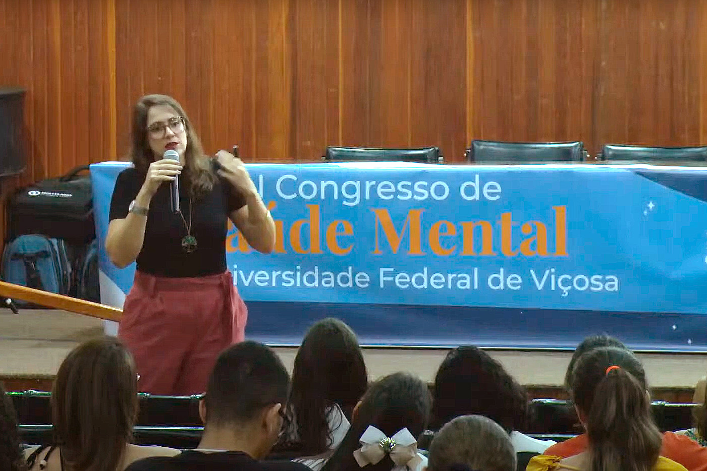 Ana Claudia está de pé em frente ao público, segurando um microfone na altura do queixo . 