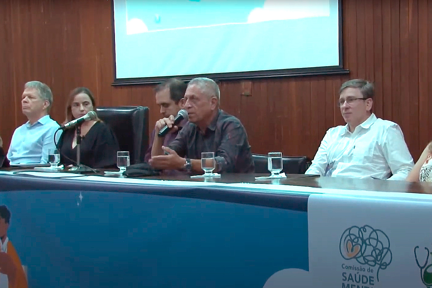 Atrás de uma longa mesa, estão 4 homens e uma mulher, todos sentados. Um dos homens segura um microfone e fala. 