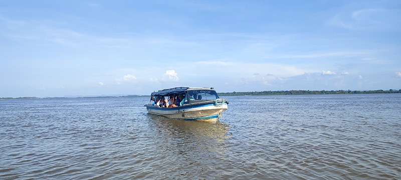 Uma pequena embarcação branca e azul navega por um rio largo de águas calmas. Ao longe, a margem formada por morros baixos. 