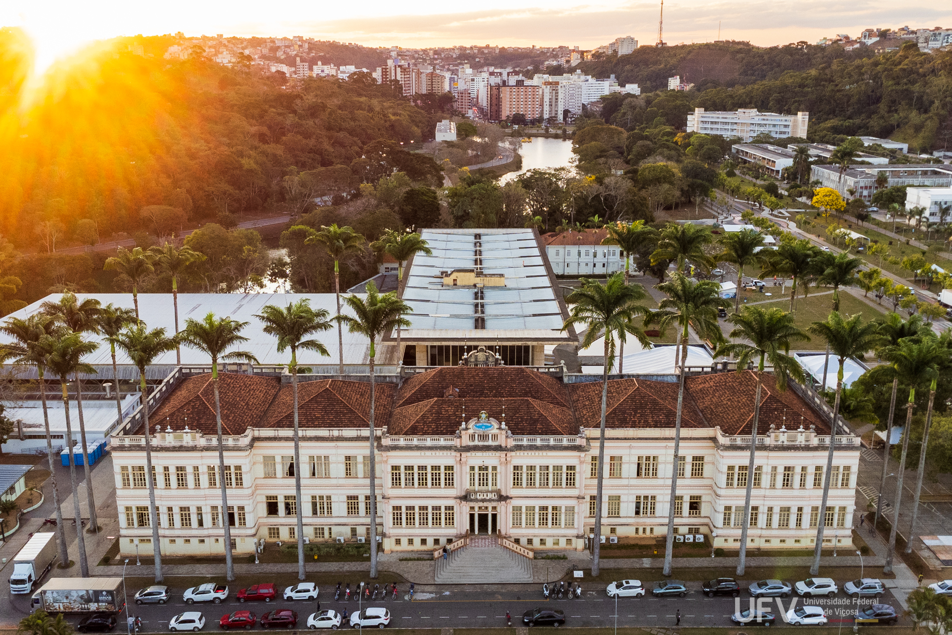 Foto aérea do pôr-do-sol mostrando prédios da universidade em primeiro plano e a cidade de Viçosa ao fundo. 