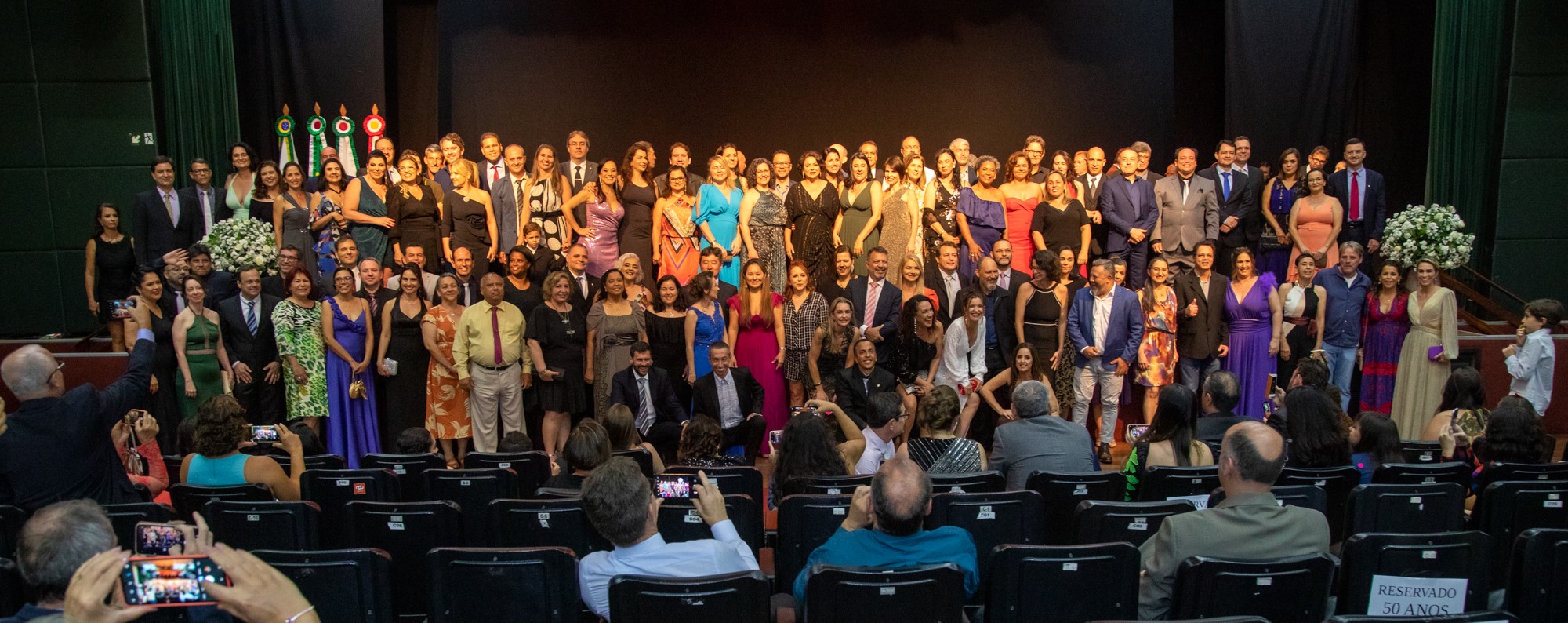 Dezenas de ex-alunos em roupas de festa posam para a foto num auditório de cortinas e cadeiras pretas. 