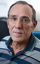 Homem de meia-idade, pele parda, magro, óculos corretivos, lábios finos e verruga no canto esquerdo da boca. 