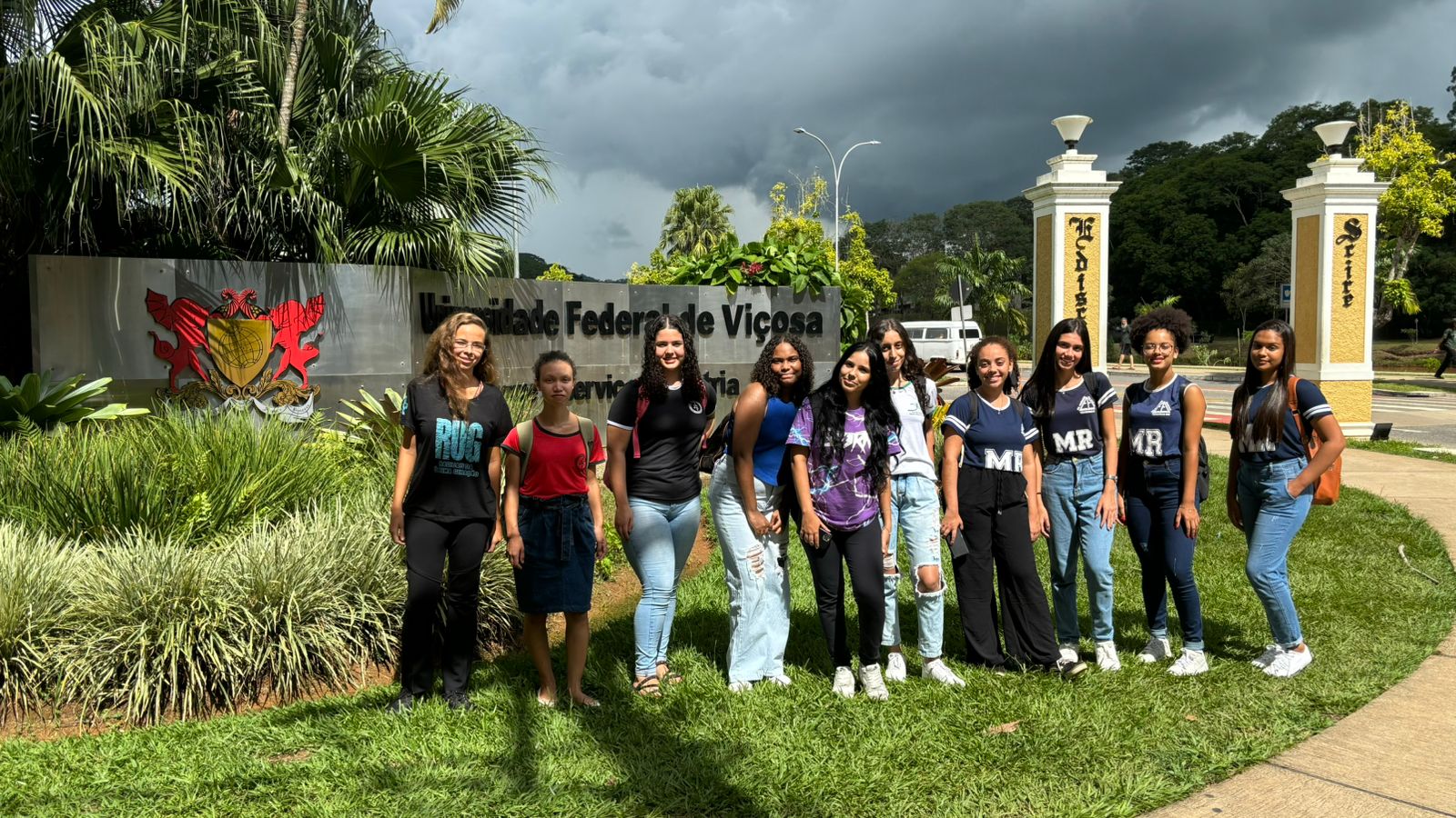 10 moças jovens de pé lado a lado posam para foto em frente a uma placa cinza com o brasão da UFV. 