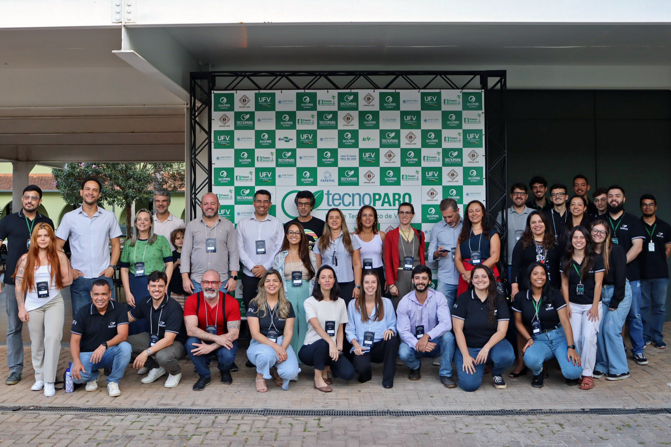 Grande grupo de pessoas de diferentes gêneros e idades, alguns de pé e outros ajoelhados, todos com crachás. 