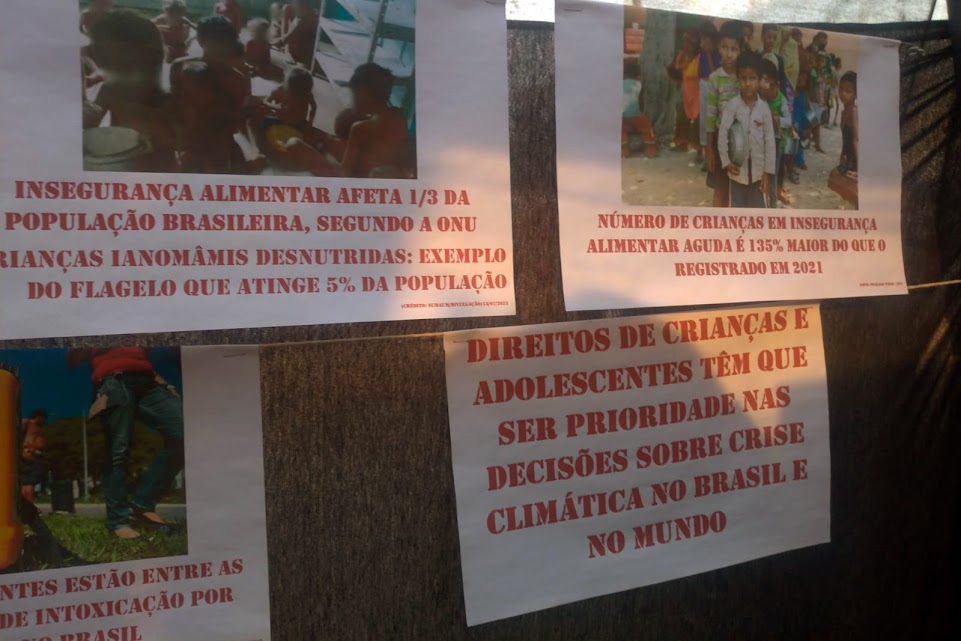 Cartazes pendurados em fios de barbante esticados,  com fotos de crianças, algumas em fila segurando pratos vazios, algumas mal vestidas se alimentando sentadas no chão. Os textos falam sobre segurança alimentar. 