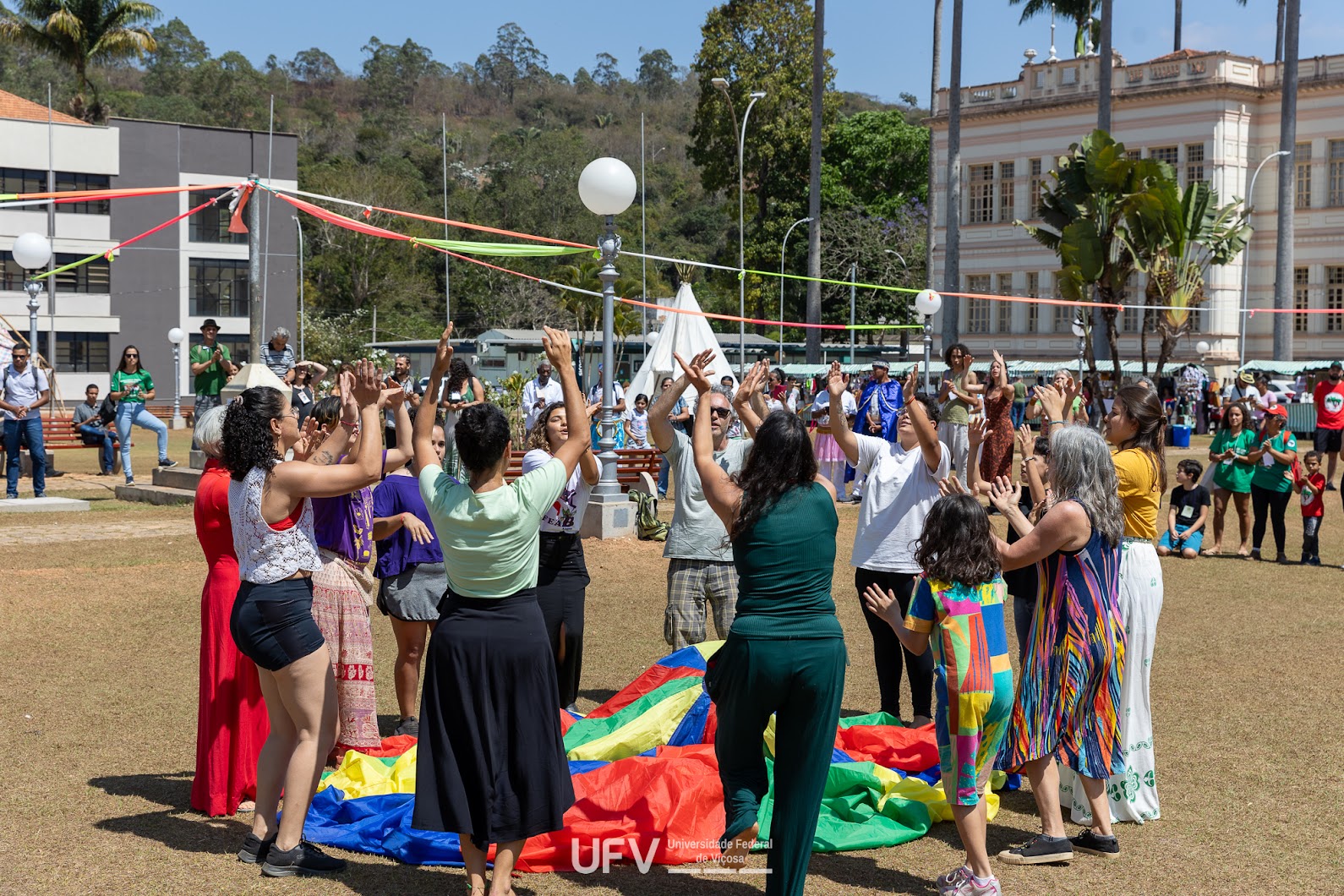 Em um grande espaço gramado, várias pessoas de várias idades e gêneros estão de pé formando um círculo e parecem cantar e dançar, com os braços erguidos acima da cabeça. Elas stão ao redor de um tecido multicolorido. 