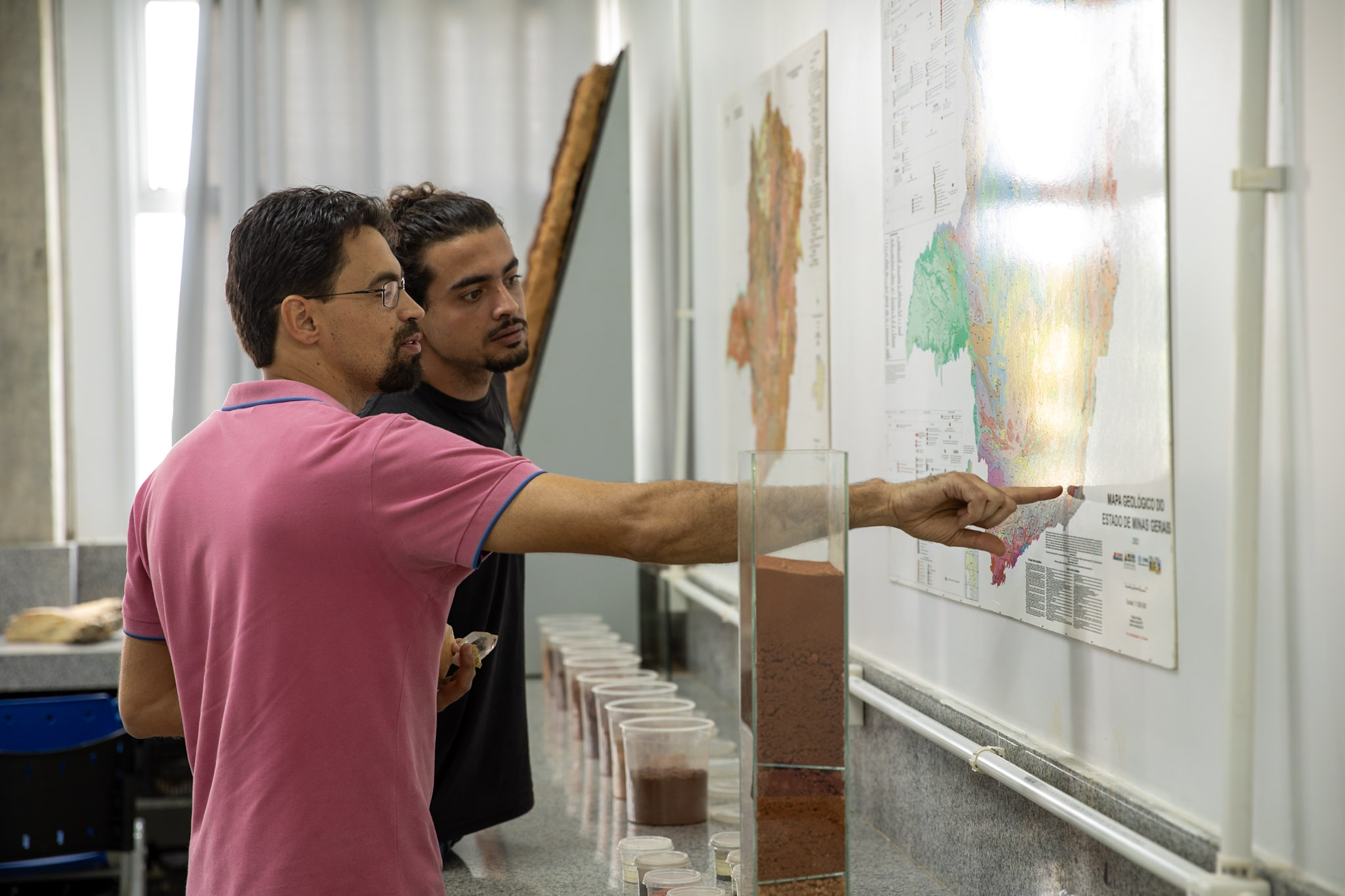 Dois homens de pé observam um mapa de Minas Gerais. Um deles está com o braço direito estendido apontando um ponto no mapa. 