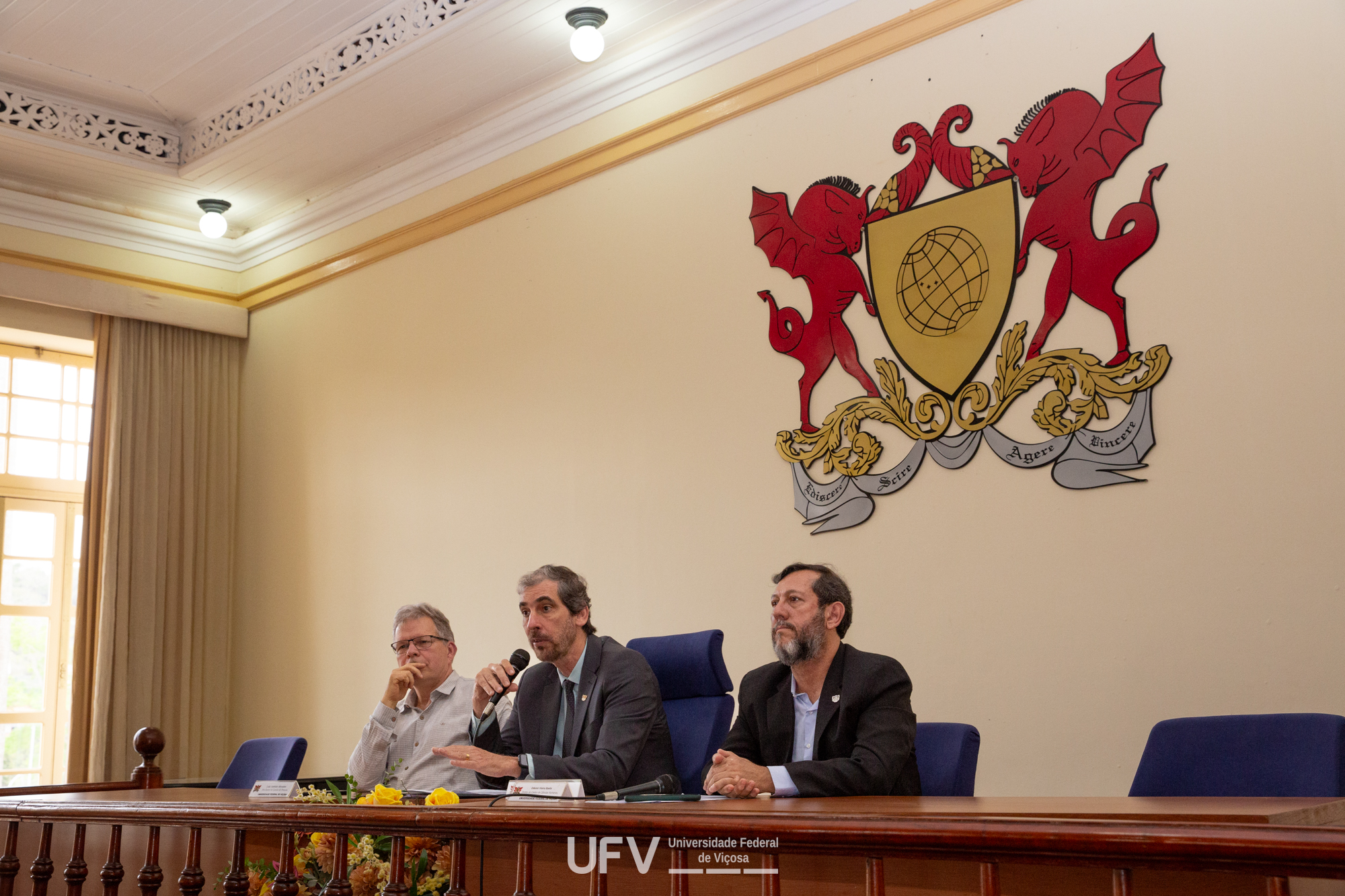 3 homens sentados  atrás de uma mesa de madeira, um deles fala ao microfone. Atrás deles, brasão da UFV numa parede bege. 