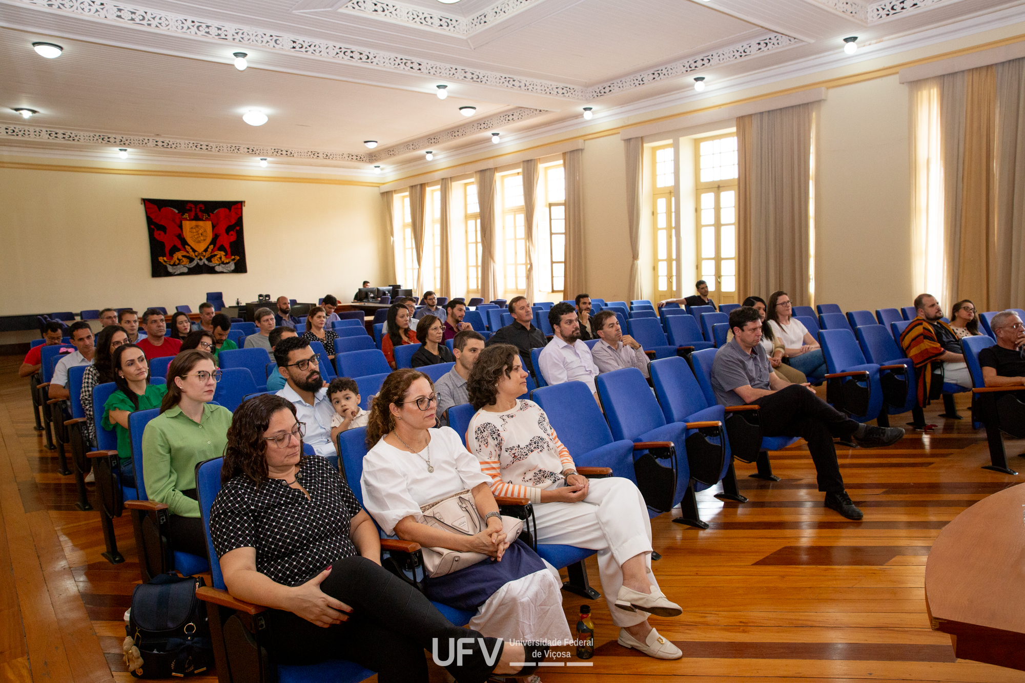 Auditório com cadeiras acolchoadas azuis, muitas delas ocupadas por homens e mulheres. 