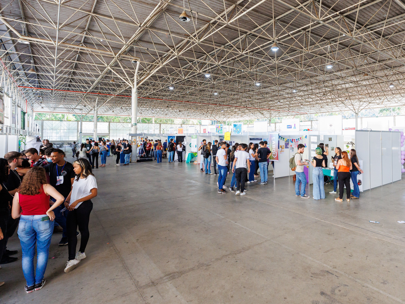 Grande espaço coberto com piso de concreto cinza. Alguns painéis brancos estão distribuídos pelo local. Dezenas de jovens estão agrupados perto destes painéis e conversam entre si. 