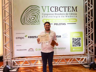 João Gilberto segura seu certificado posando para a foto em frente a um painel  com o texto quinto C B C TEM. 