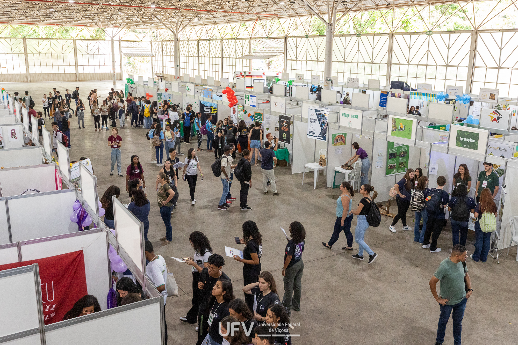 Grande espaço coberto com stands brancos espalhados. Muitos jovens circulam pelo ambiente. 