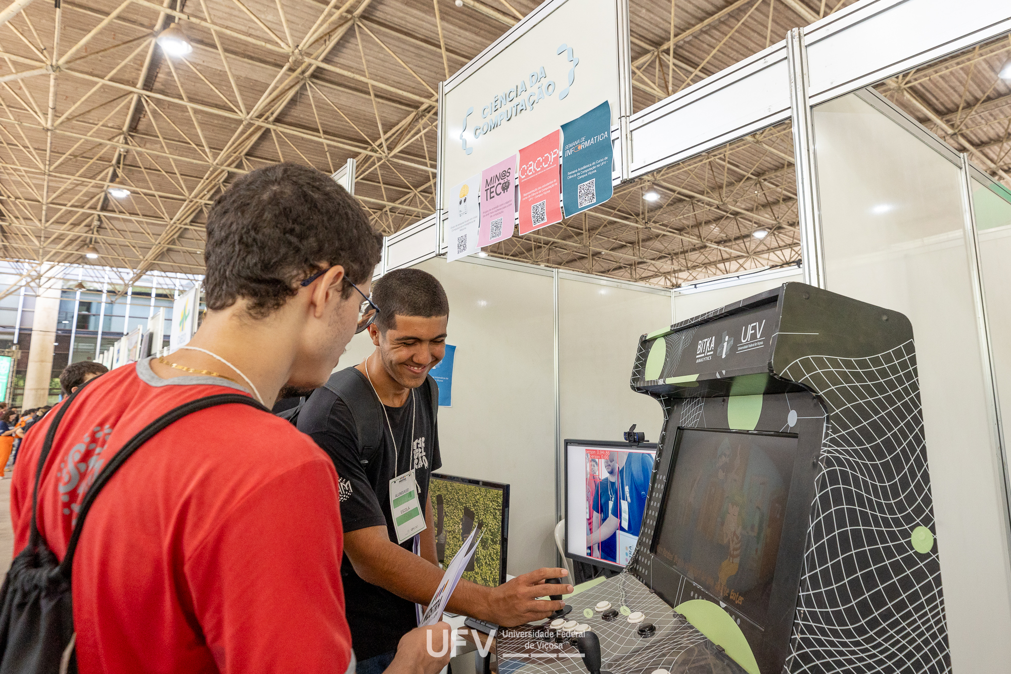 Dois jovens manipulam um videogame tipo fliperama, dentro de um dos stands do evento. 