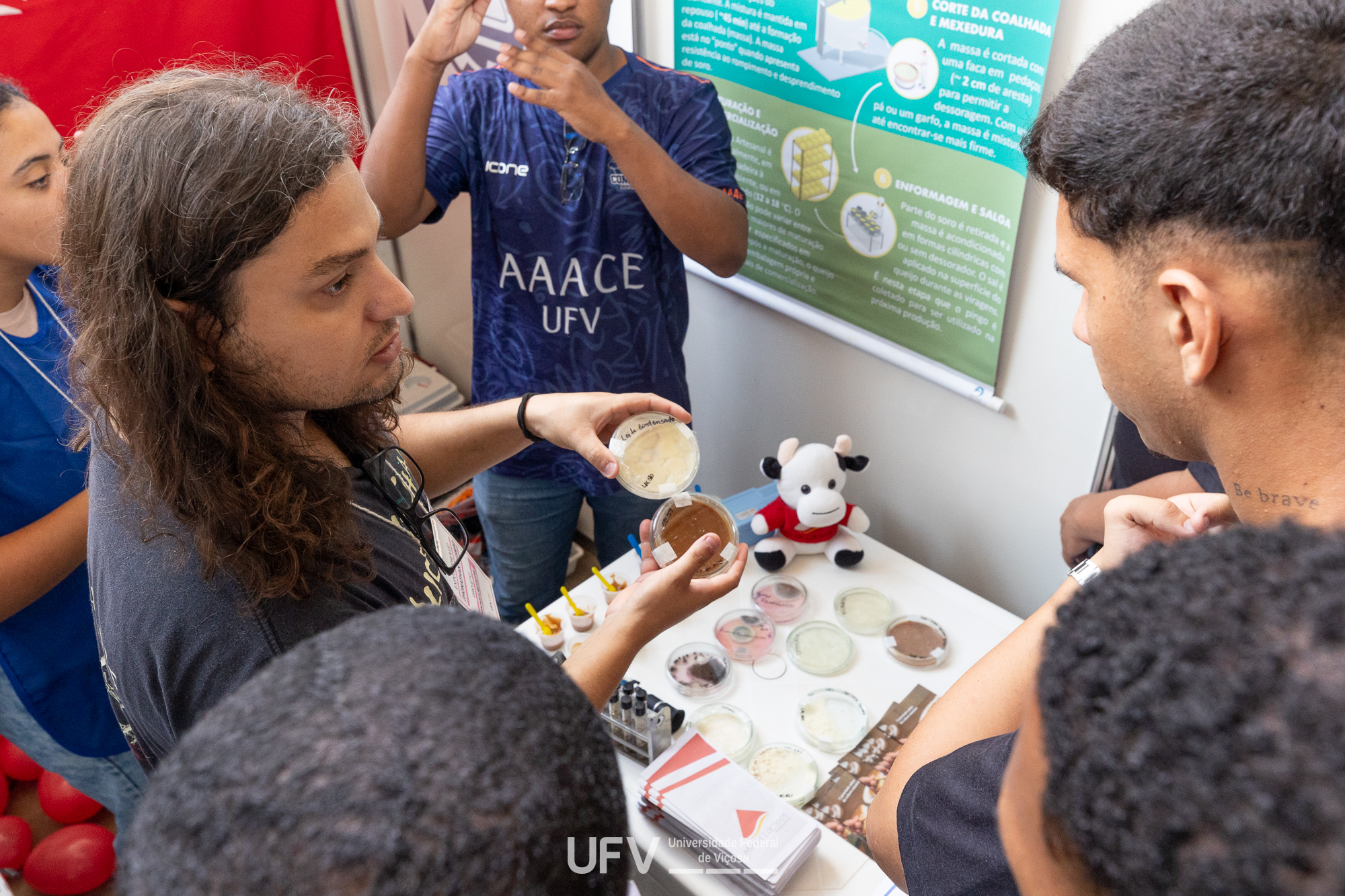 Um jovem de cabelos longos e ondulados mostra placas de cultura para outros jovens. 