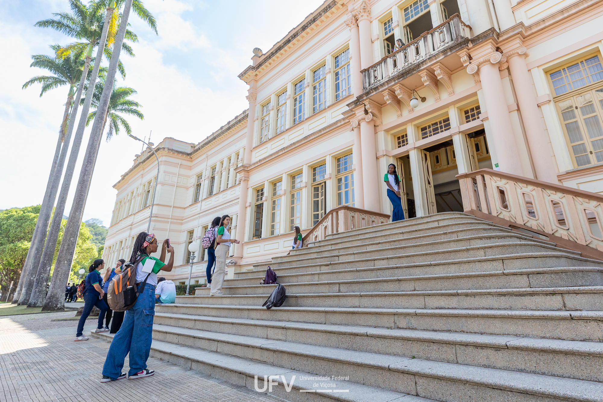 Algumas jovens na escadaria de um edifício de arquitetura antiga. Uma delas posa para que outra a fotografe. 