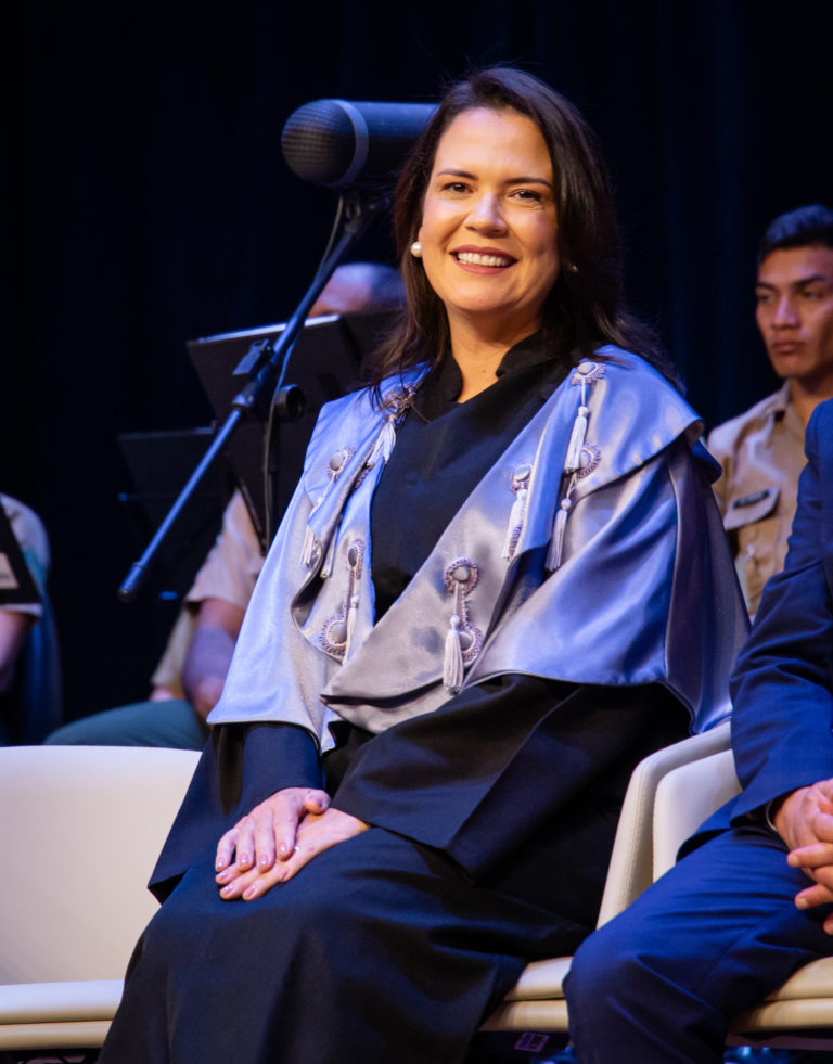 Mulher branca, cabelos lisos castanhos e longos, usa beca preta e pelerine azul clara com adornos de linha. Ela sorri.