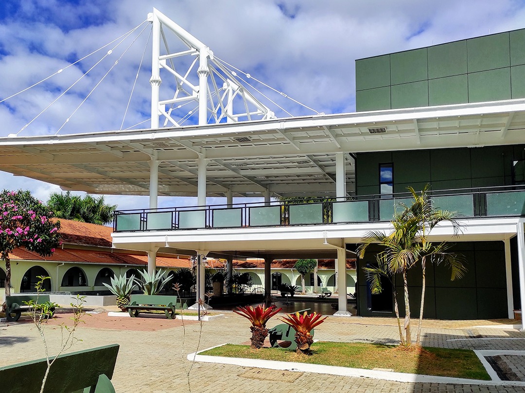 Edifício baixo verde, com uma grande varanda cuja cobertura é sustentada por hastes e pilares metálicos brancos.  